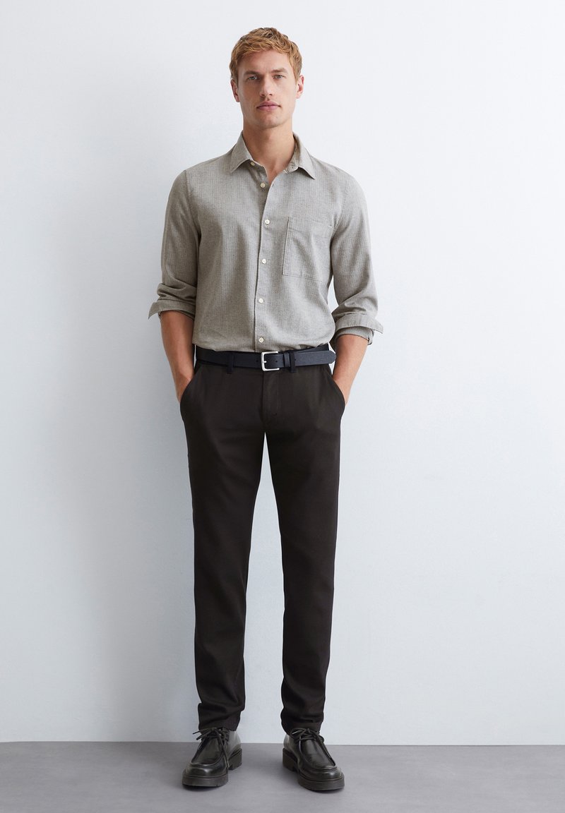 Helles graues Hemd mit Knopfleiste und Tasche; schwarze Hose; schwarze Schuhe; lockere Passform; hochgekrempelte Ärmel, minimalistisches Design, keine Muster.