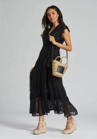 Robe noire à manches volantées, avec des couches transparentes et des embellissements subtils. Assortie à un sac en cuir tressé et des sandales compensées.