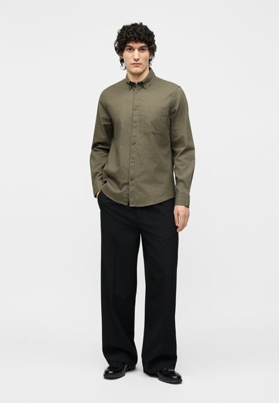 Jeune homme debout les mains dans les poches portant une chemise boutonnée vert olive, un pantalon noir à jambes larges et des chaussures noires sur un fond uni.