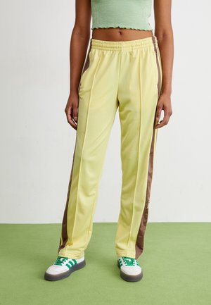 Persoon draagt lichtgele trainingsbroek met bruine zijstrepen, een cropped groene top en witte sneakers met groene strepen, staande op een groene vloer.