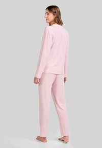 Mujer de pie descalza, girada ligeramente de perfil, vistiendo una blusa de manga larga rosa suave y pantalones a juego, frente a un fondo gris liso.