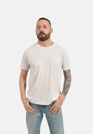 Uomo con barba e orecchini che indossa una semplice maglietta beige chiaro e jeans blu strappati, mostrando una manica colorata di tatuaggi sul braccio destro.