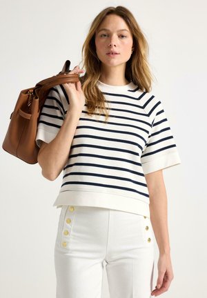 Kvinde iført en hvid og navystribet kortærmet top og hvide bukser med guldknapper, bærende en brun lædertaske over skulderen.