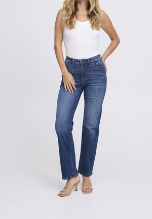 Femme portant un haut blanc sans manches, un jean bleu à jambes droites et des talons ouverts beige, debout avec une main sur la hanche.