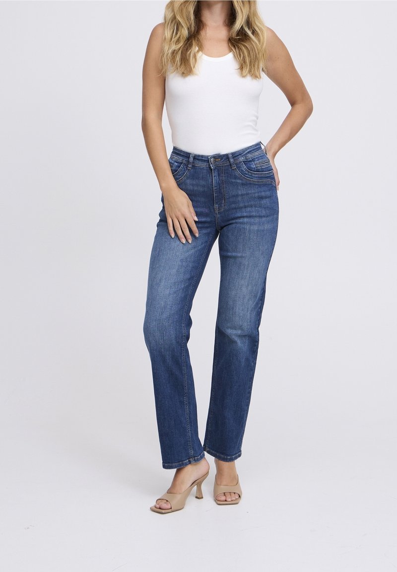 Femme portant un haut blanc sans manches, un jean bleu à jambes droites et des talons ouverts beige, debout avec une main sur la hanche.