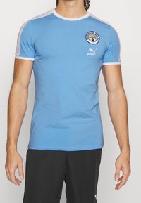 Muž oblečený v světle modrém tričku Manchester City FC od Pumy a černých šortkách od Pumy, stojící proti bílému pozadí.