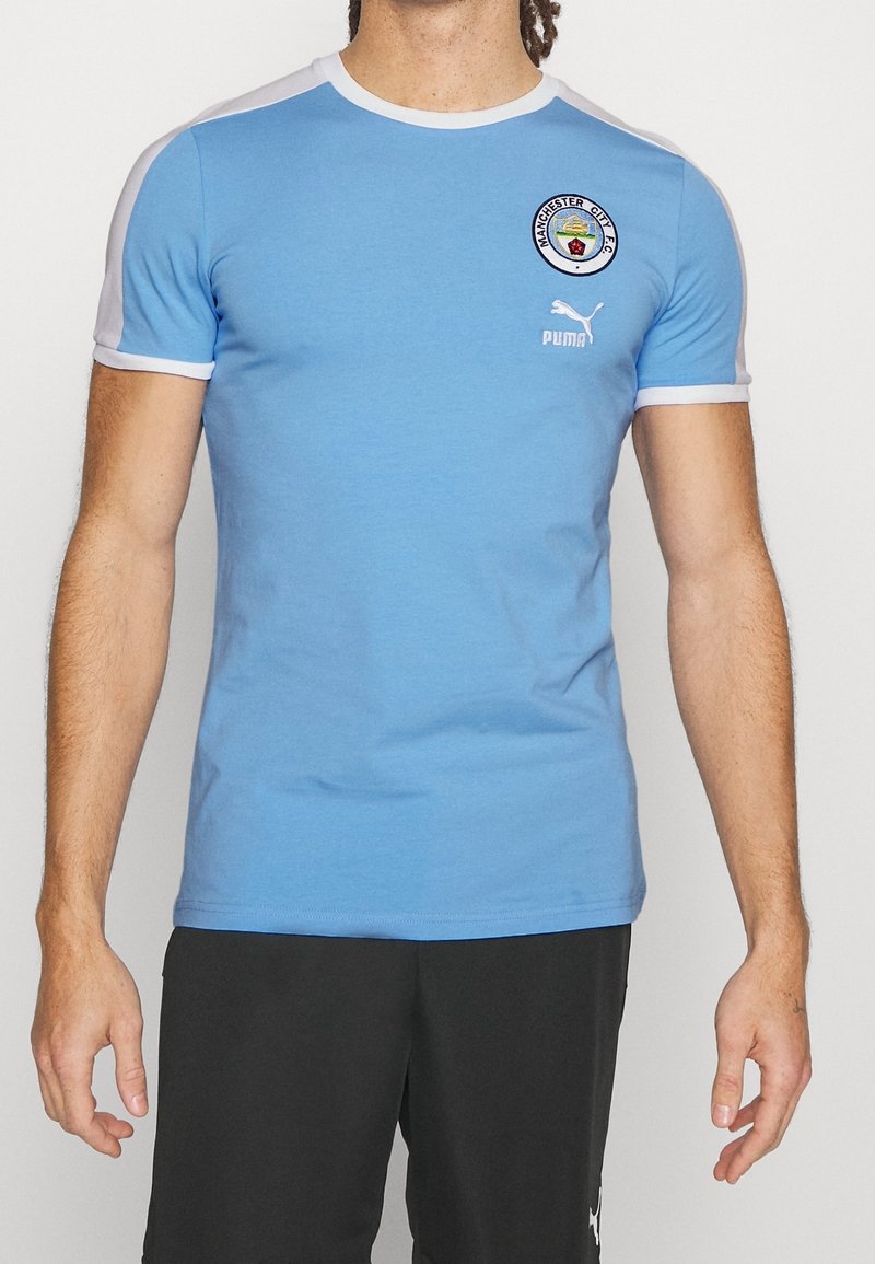 Muž oblečený v světle modrém tričku Manchester City FC od Pumy a černých šortkách od Pumy, stojící proti bílému pozadí.