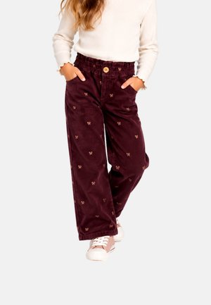 Enfant portant un haut crème à manches longues et un pantalon large violet foncé avec de petits motifs de nœuds, mains dans les poches, et des baskets roses.