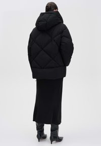 Doudoune noire avec capuche, présentant un motif en losange et un design court, associée à une longue jupe noire et des bottes jusqu'aux genoux.