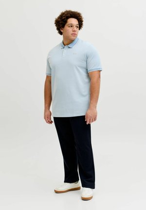 Lyseblå poloshirt med en flad krave, korte ærmer og diskret logo. Kombineret med mørke bukser og hvide sneakers, hvilket skaber et afslappet look.