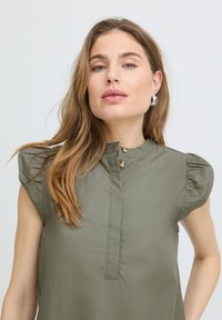 Jeune femme aux longs cheveux châtain clair portant des boucles d'oreilles anneaux en argent et un chemisier sans manches vert olive avec des épaules bouffantes.