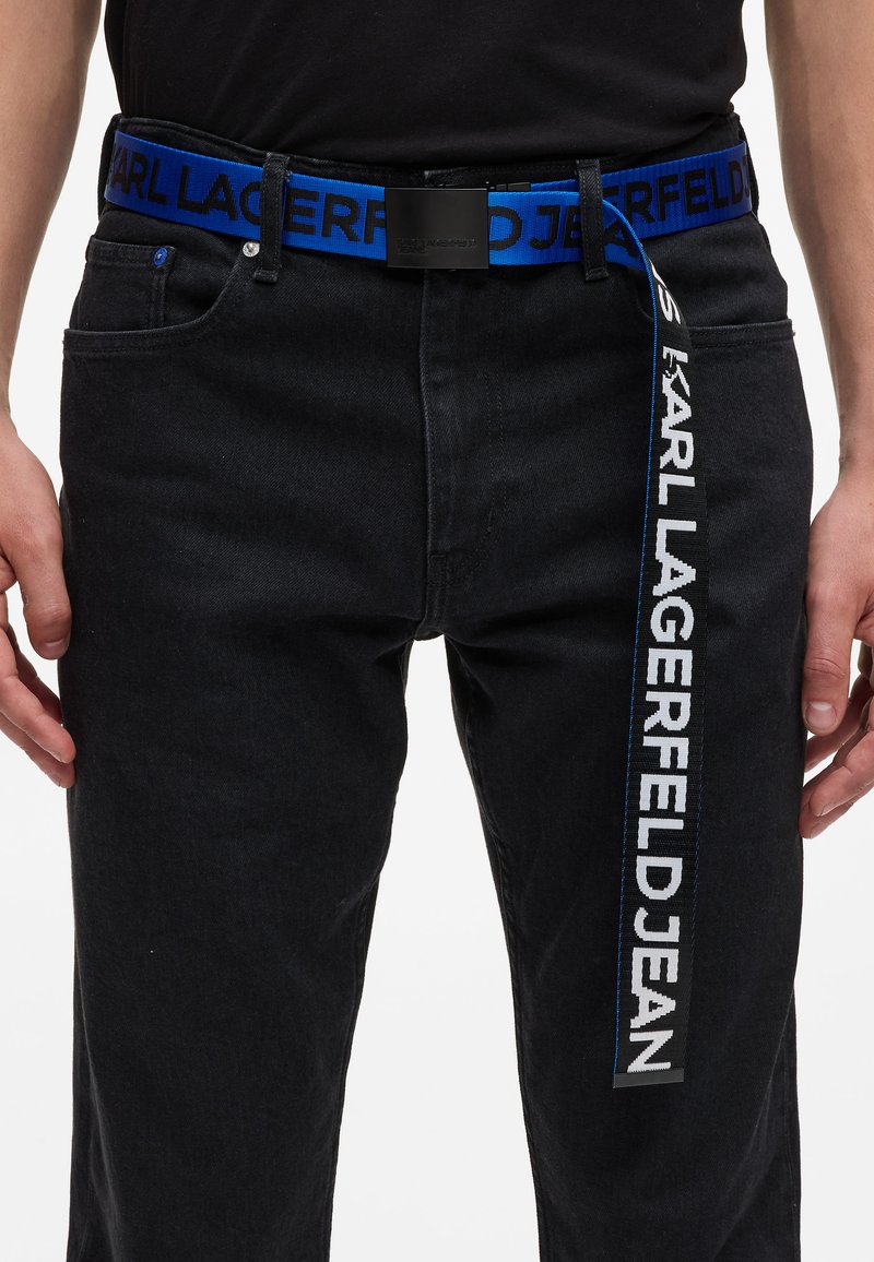 Kék pamut öv fekete betűkkel: "KARL LAGERFELD JEAN." Fém csat. Fekete farmernadrág fölött viselve.