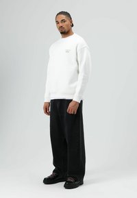 Witte sweater met geribbelde manchetten en zoom, voorzien van een klein geborduurd logo op de borst, gecombineerd met losse zwarte denim broek en zwarte schoenen.