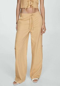 Pantalones anchos, ligeros y beiges con cintura de cordón y bolsillos laterales, hechos de una tela suave con una textura lisa.
