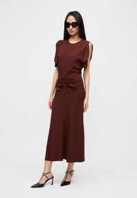 Μακρύ μίντι φόρεμα σε μπορντό με λεπτομέρειες συγκέντρωσης στη μέση, κοντά φουσκωτά μανίκια και ομαλή υφή, συνδυασμένο με μαύρα παπούτσια με τακούνι.