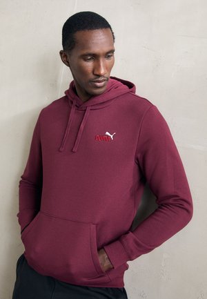 Man in een kastanjebruine Puma hoodie met zijn handen in de voorzak, kijkend naar beneden, staand tegen een lichte, gestructureerde muur.