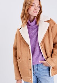 Giacca in suede marrone con colletto in eco-pelliccia bianca, con chiusura a bottoni e tasche laterali, indossata sopra un dolcevita viola e jeans blu.