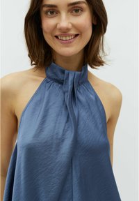 Zila halter tipa blūze ar pleitu dizainu, kas izceļas ar gludu, spīdīgu tekstūru un atvērtu muguru, izrādot modeļa plecus.