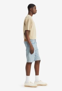 Beige cotton t-shirt, light blue denim shorts, white socks, and cream sneakers. Model stands sideways against a plain white background.