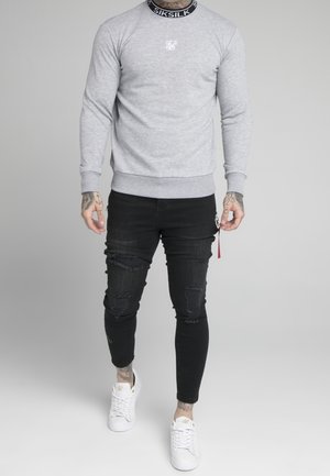 Sudadera gris con cuello de canalé, que presenta un pequeño logo blanco. Combinada con jeans ajustados negros desgastados y zapatillas blancas.