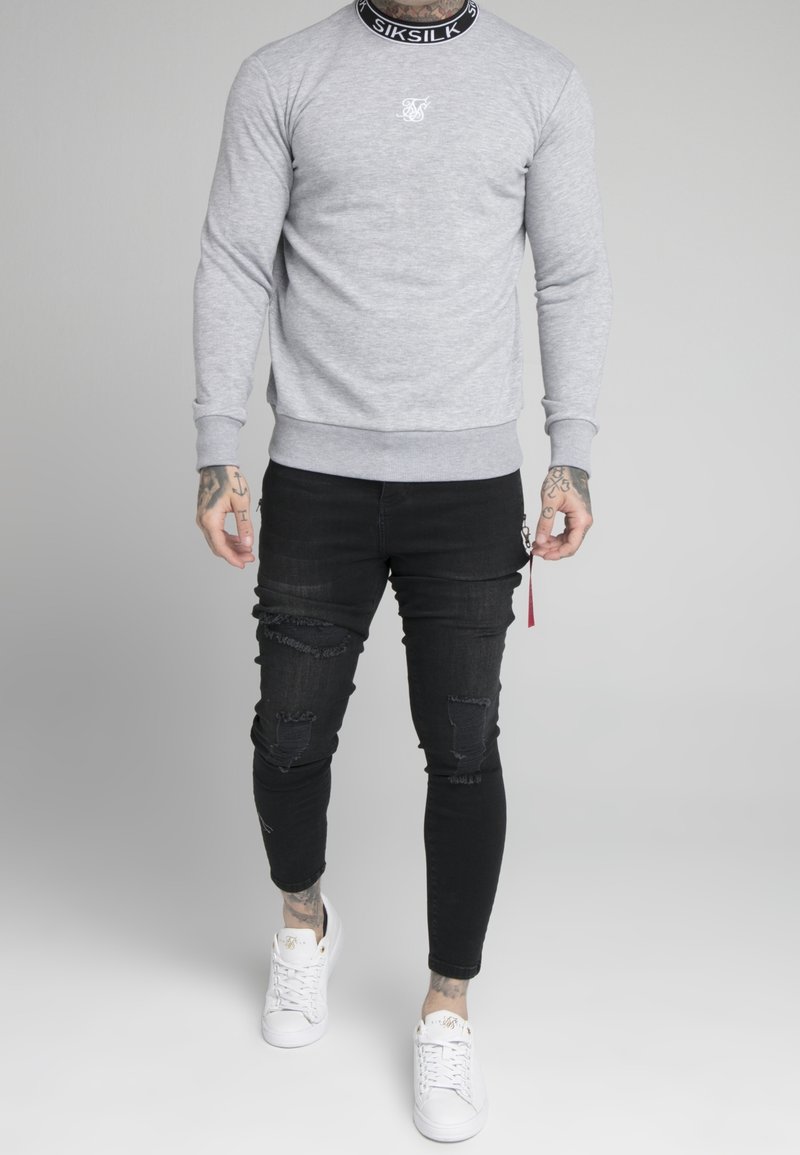 Grå sweatshirt med ribbad krage, med en liten vit logotyp. Matchad med svarta slitna slim-fit jeans och vita sneakers.