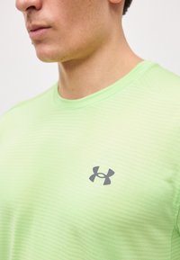 Άνδρας που φοράει αθλητική μπλούζα ανοιχτού πράσινου χρώματος με υφή και γκρι λογότυπο Under Armour στο στήθος, εμφανιζόμενος από τον λαιμό έως το μέσο του κορμού.