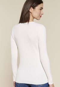 Maglione a maniche lunghe a coste di colore bianco, con un design aderente, scollatura rotonda e un orlo posteriore più lungo, indossato da un modello visto da dietro.