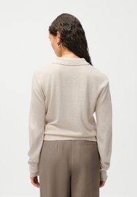 Sweater de malha bege com gola, mangas longas e bainha canelada. Usado sobre calças castanhas claras, apresentando um ajuste relaxado.