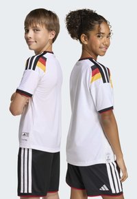 White soccer jerseys with black and red accents, paired with black shorts featuring three white stripes. Breathable textured fabric.