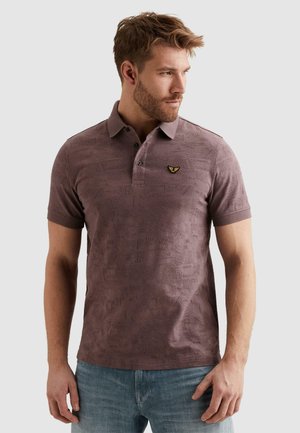 Mężczyzna o krótkich włosach i brodzie, ubrany w mauve polo z wzorem oraz jasnoniebieskie dżinsy, spogląda w prawo na jednolitym tle.