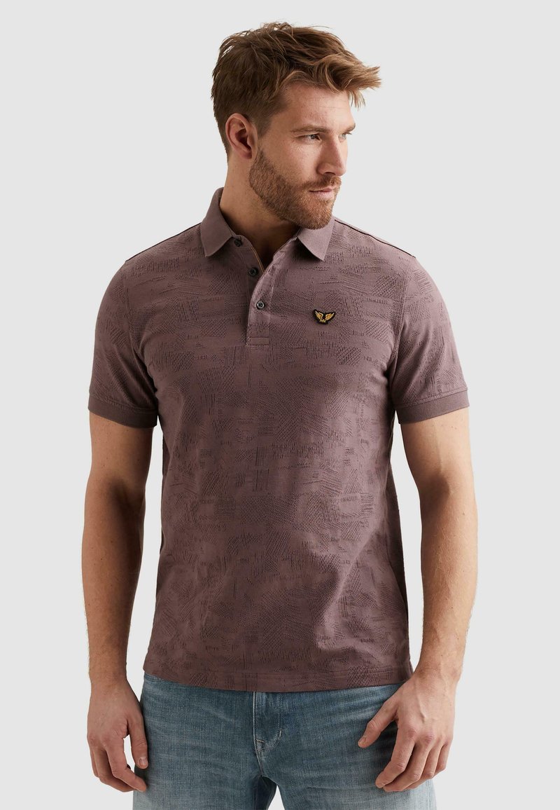 Homme aux cheveux courts et barbe, portant un polo mauve à motifs et un jean bleu clair, regardant vers sa droite sur un fond uni.