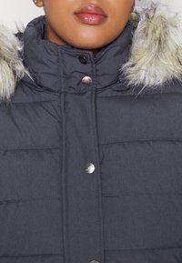 Giacca puffer grigio scuro con colletto alto, cappuccio foderato in pelliccia e bottoni a pressione argentati. Presenta una texture a trapuntatura e una finitura in tessuto liscia.