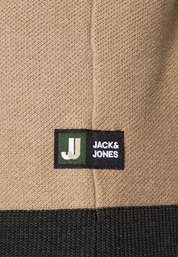 Tessuto a maglia beige con orlo a coste verde. Etichetta nera con testo bianco e verde "JACK & JONES" e logo che mostra il design "JJ".