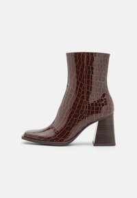 Bottine en faux cuir brun avec une texture crocodile, bout carré et un talon en bois épais. Fini lisse, silhouette épurée.