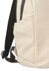 Mochila beige con correas de malla negras, forma curva, textura suave, que cuenta con un bolsillo lateral y detalles de cremallera en contraste.