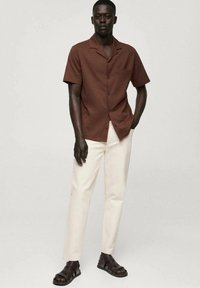 Camicetta marrone a maniche corte con colletto, abbinata a pantaloni crema e sandali marroni. La camicia presenta bottoni e una tasca sul petto.