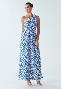 Maxi haljina plavo-bijelog zebra uzorka, s jednim ramenom, strukiranim strukom i lepršavim materijalom. Nošena s bijelim sandalama s remenčićima.