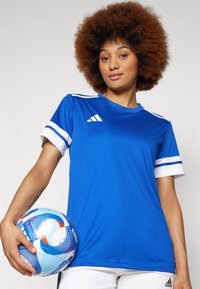 Camisola desportiva azul com detalhes em faixa branca, mangas curtas e gola redonda. A segurar uma bola de futebol com padrão, exibida contra um fundo neutro.