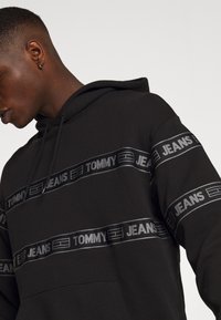 Felpa nera con cappuccio, tasca a marsupio, coulisse per il cappuccio e polsini elasticizzati. Presenta strisce orizzontali con il logo "TOMMY JEANS" in grigio.
