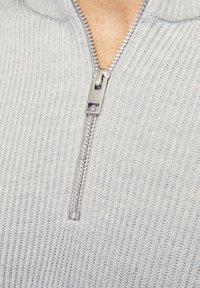 Suéter gris claro con alto cuello y cremallera plateada. Presenta un textura acanalada, costuras visibles y un diseño ajustado.