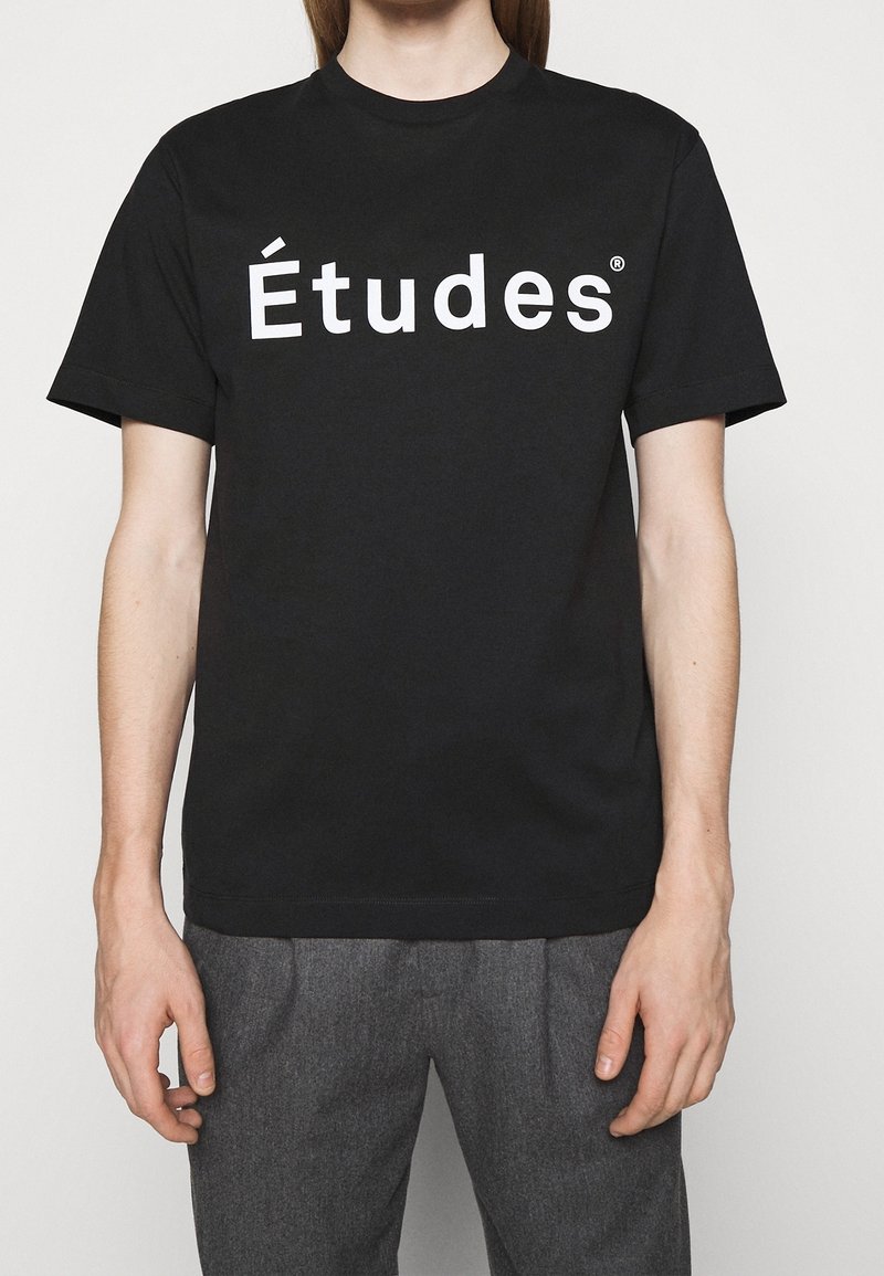 Personne portant un T-shirt noir avec le texte blanc "Études" sur la poitrine et un pantalon gris, se tenant devant un fond clair.