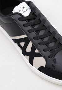 Zapatilla de cuero negra con un diseño texturizado en negro y beige, que presenta un prominente patrón de cortes geométricos y cordones negros.