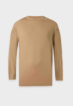 Jeune homme portant une chemise marron à manches longues, un jean bleu clair à jambes larges et des chaussures noires, debout devant un fond blanc uni.