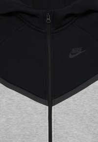 Nike zip-up hoodie featuring a black upper, grey lower panel, and contrasting grey accents. Material is smooth with a subtle texture.