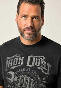 Camiseta de algodón negra con un diseño gráfico que incluye el texto "Iron Dust" y una mano esquelética sosteniendo una llave inglesa, con acentos plateados desgastados.