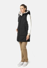 Chaleco acolchado negro con capucha, que cuenta con dos bolsillos laterales, una cremallera frontal y un forro interior beige. Lleva puesto sobre pantalones beige claros y zapatillas blancas.
