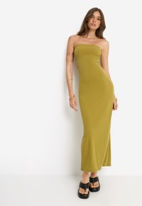 Μάξι φόρεμα σε ελιά πράσινο, ριγωτό, χωρίς τιράντες, με εφαρμοστή σιλουέτα. Συνδυασμένο με χοντρούς μαύρους σανδάλια, που αναδεικνύουν ένα λείο ύφασμα.