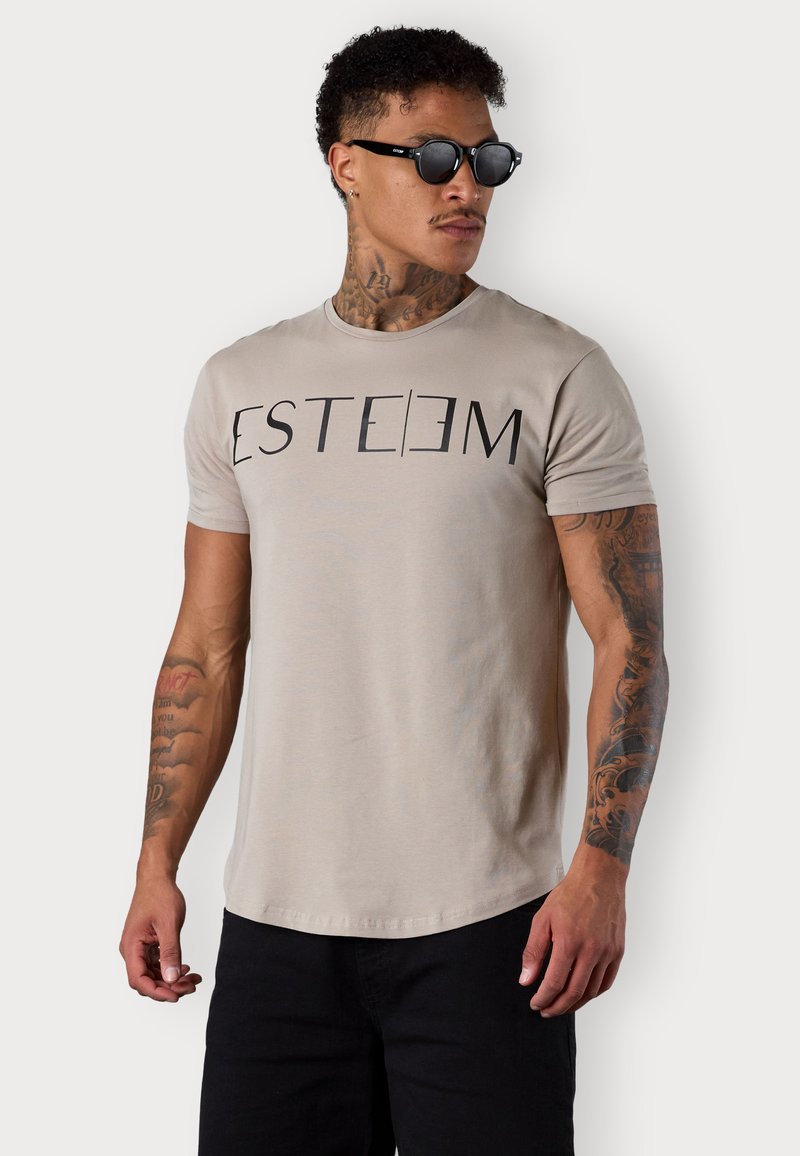 T-shirt en coton beige avec le texte noir "ESTEEM", col rond, manches courtes et coupe décontractée. Le modèle porte un short foncé et des lunettes de soleil noires.