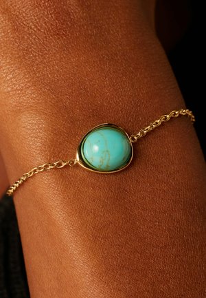 Goldene Kettenarmband mit einem runden, türkisen Stein mit goldenen Aderungen, der in einer glatten Fassung sitzt, vor einem strukturierten Hauthintergrund.