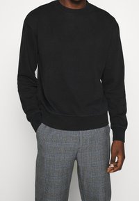 Sudadera negra con cuello redondo, mangas largas y puños acanalados, combinada con pantalones de cuadros grises con acentos azules.